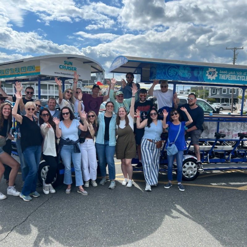 Group of people posing happily in front of a large pedal-powered vehicle.