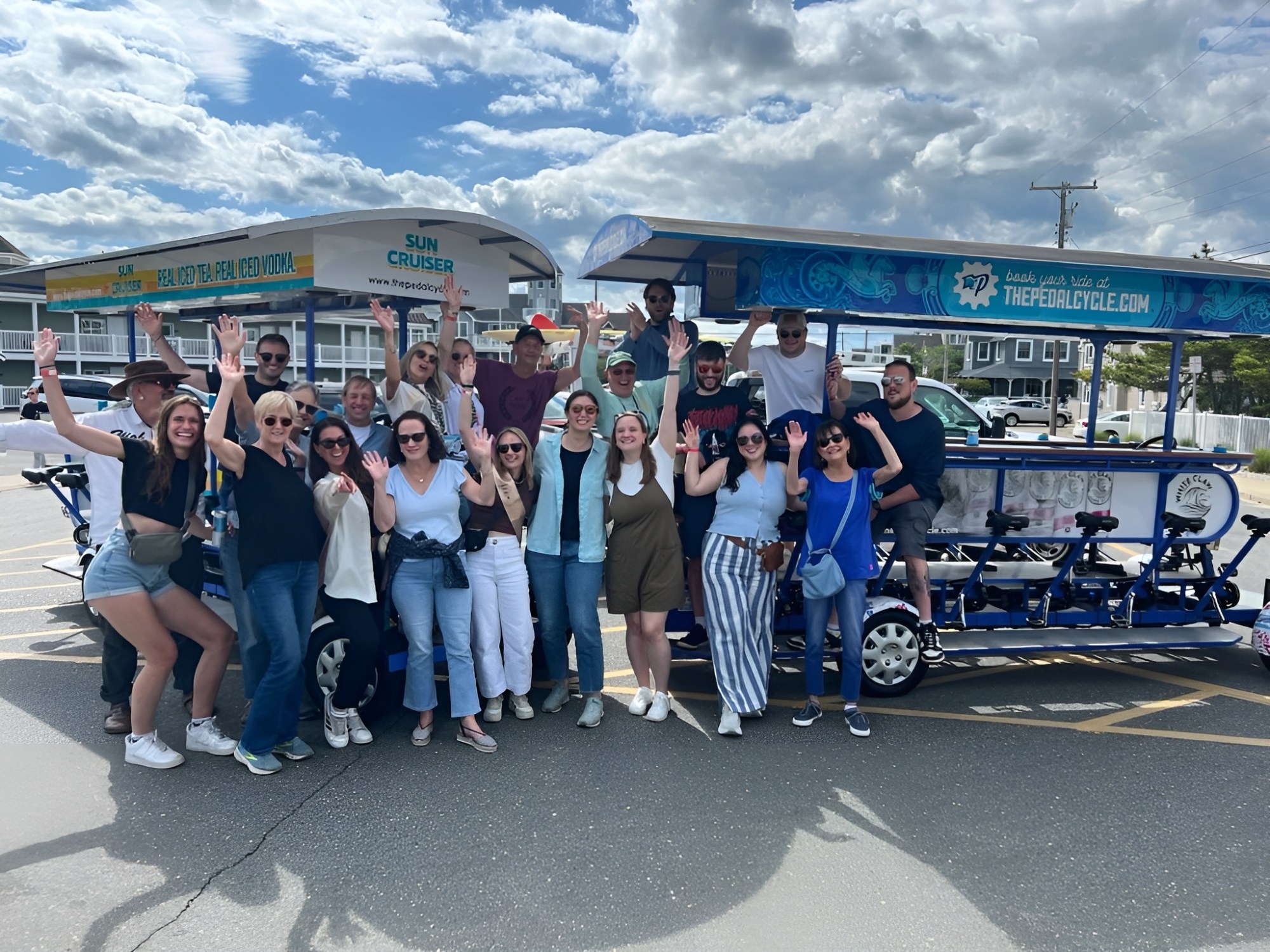 Group of people smiling and waving, standing around a blue pedal cycle on a sunny day.