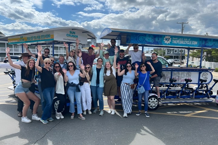 Group of people smiling and waving, standing around a blue pedal cycle on a sunny day.