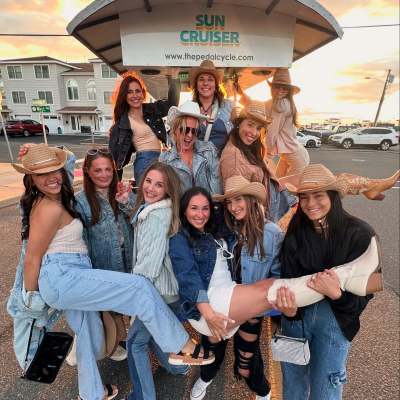 Group of people in cowboy hats and denim outfits posing happily beside a Sun Cruiser with sunset background.