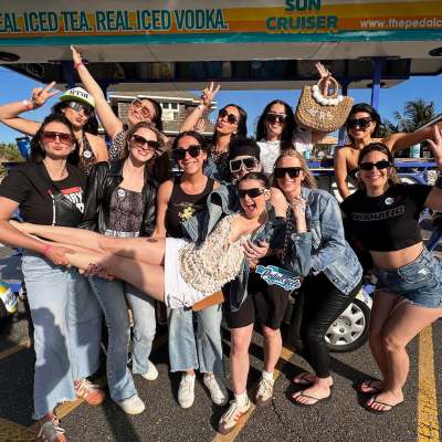 Group of women posing playfully outdoors, holding one in the air, with sunglasses and a pedal cart in background.