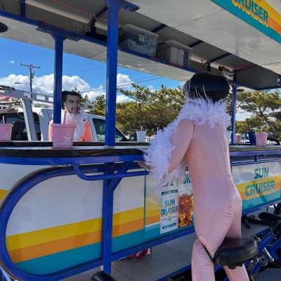 Person in pink suit and boa on a pedal bar with Sun Cruiser sign.