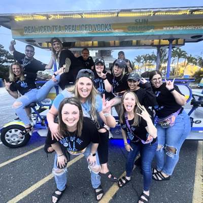 Group of people on a pedal-powered vehicle posing cheerfully for a photo, many making rock-on hand signs.