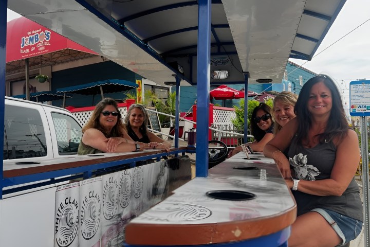 Five women sitting at an outdoor bar counter, smiling with a restaurant in the background.
