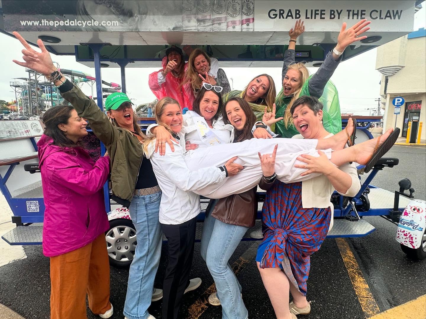 Group of women smiling and holding one person horizontally in celebration near a pedal cycle.
