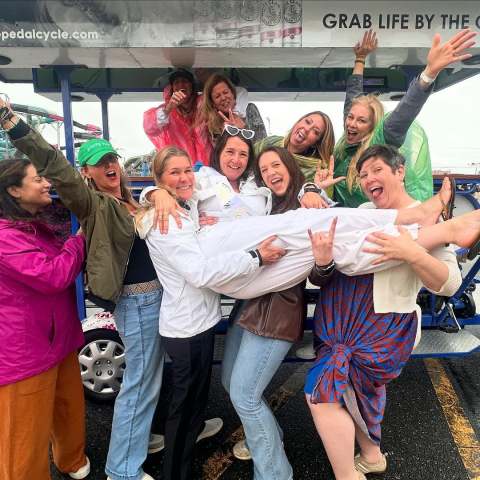 Group of women smiling and holding one person horizontally in celebration near a pedal cycle.