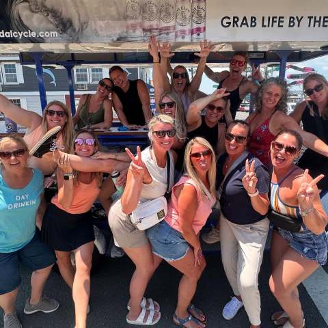 Group of women posing energetically in front of a pedal-powered vehicle on a sunny day.
