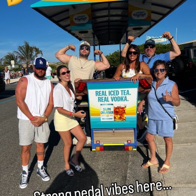 Group posing in front of pedal cycle with flexed arms, sign reading 'Real Iced Tea. Real Vodka.'