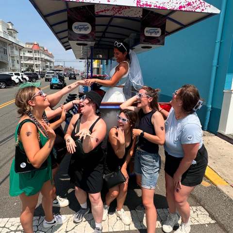 Bride in cart with veil and sunglasses laughing with six women on the street.