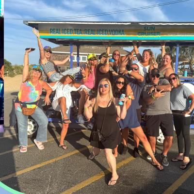 Group of people posing playfully in front of a sign for iced tea and vodka on a sunny day.