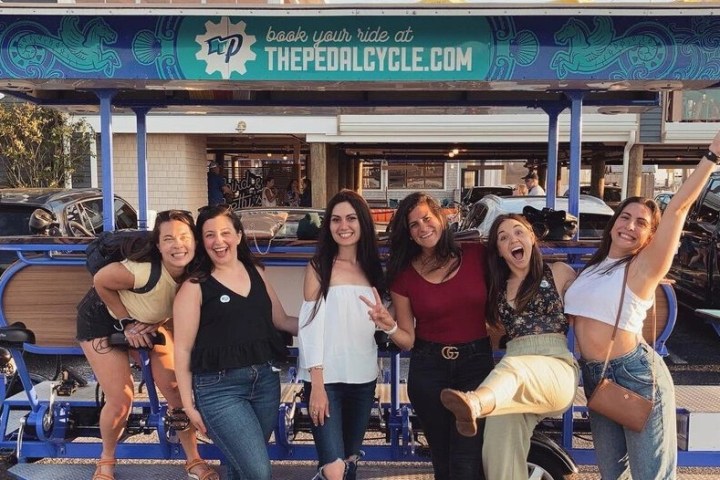 Six women smiling and posing in front of a pedal vehicle in a parking lot.
