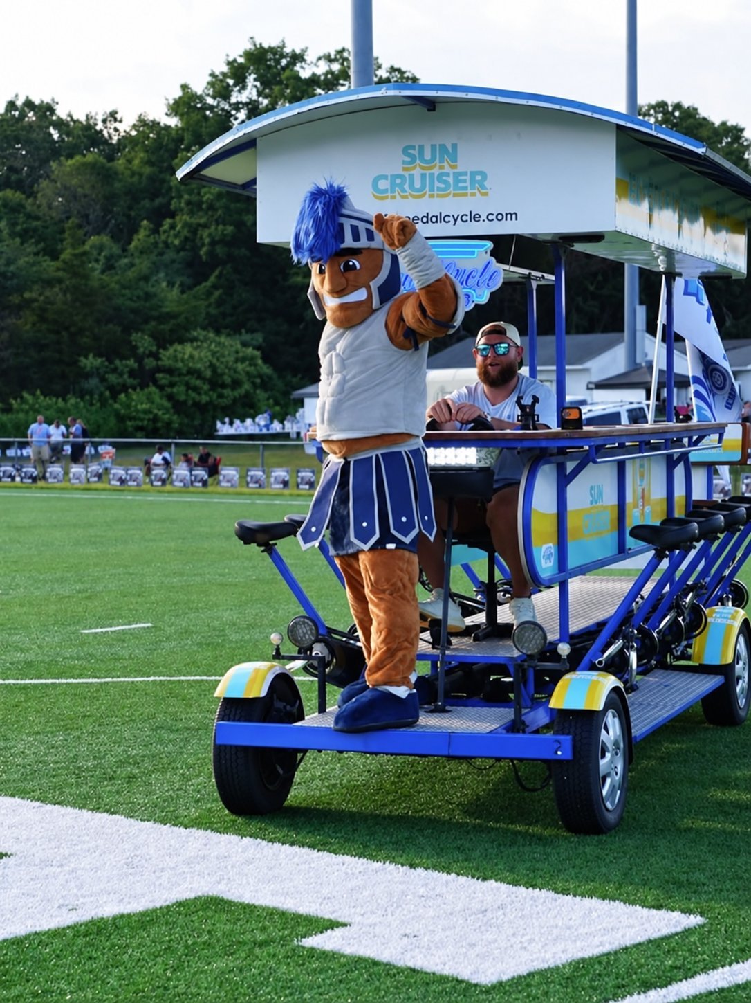 Mascot on pedal-powered vehicle at sports field.